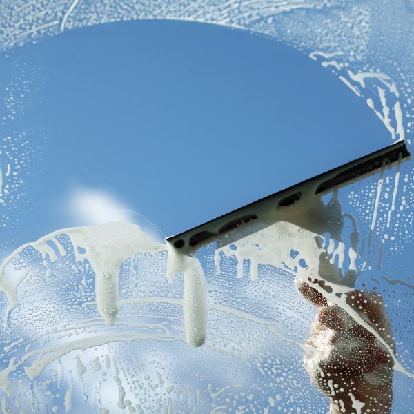 A hand cleaning a soapy window with a squeegee against a clear blue sky.