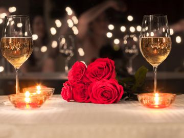 Two filled white wine glasses on a table, with red roses, and lit tealights.