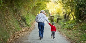 Older man and child holding hands, walking on trail with trees on either side. 