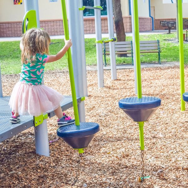 A young girl in a pink tutu plays on green and blue playground equipment outdoors.
