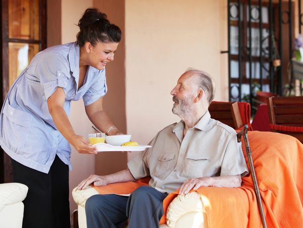 Caregiver serving breakfast to elderly man in armchair.