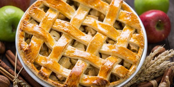 Golden lattice-top apple pie surrounded by fresh apples and cinnamon sticks.