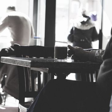 A quiet cafe scene with a person holding a cup and blurred figures outside.