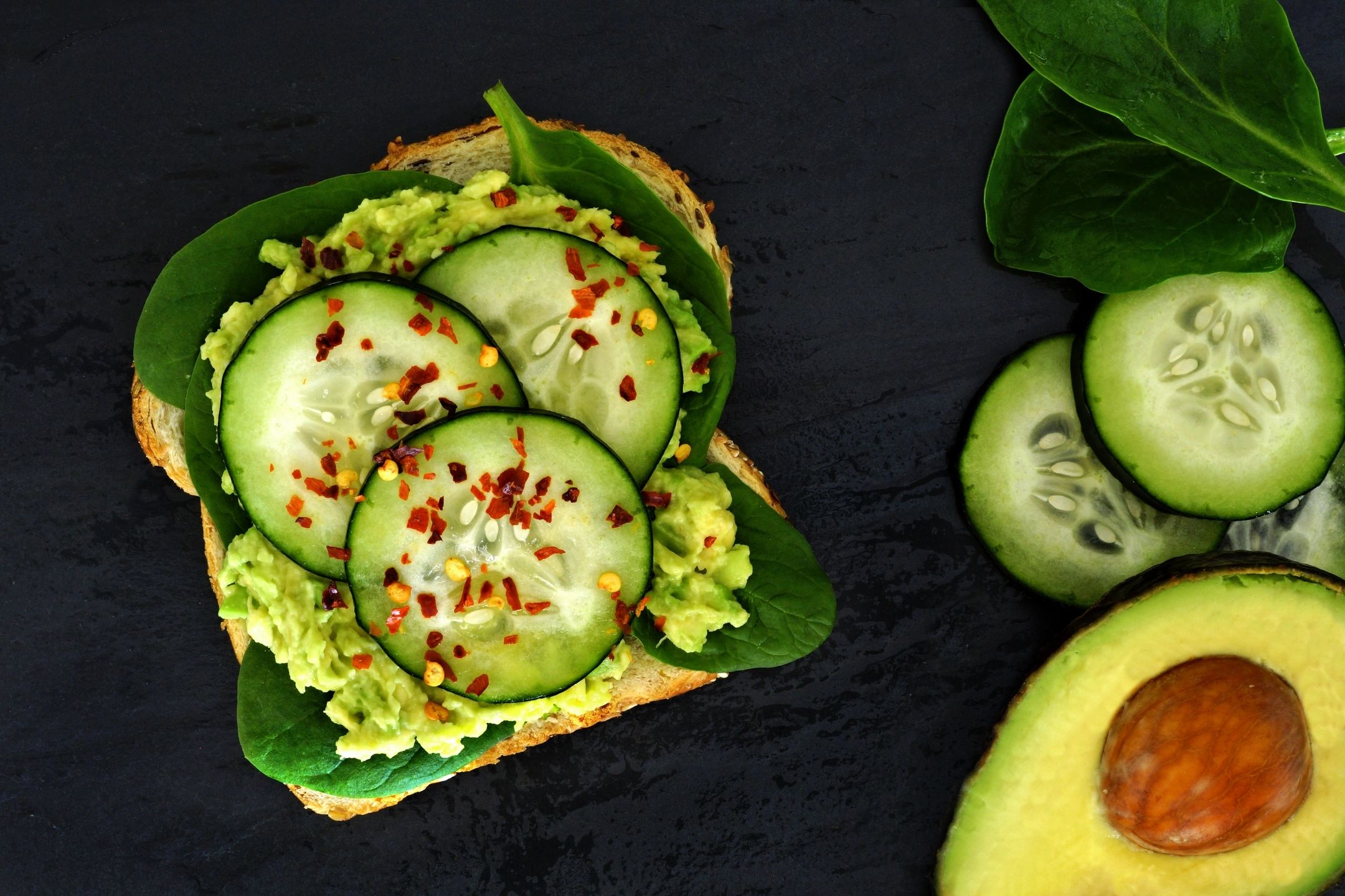 Avocado toast with cucumber slices, spinach, and chili flakes on dark background.