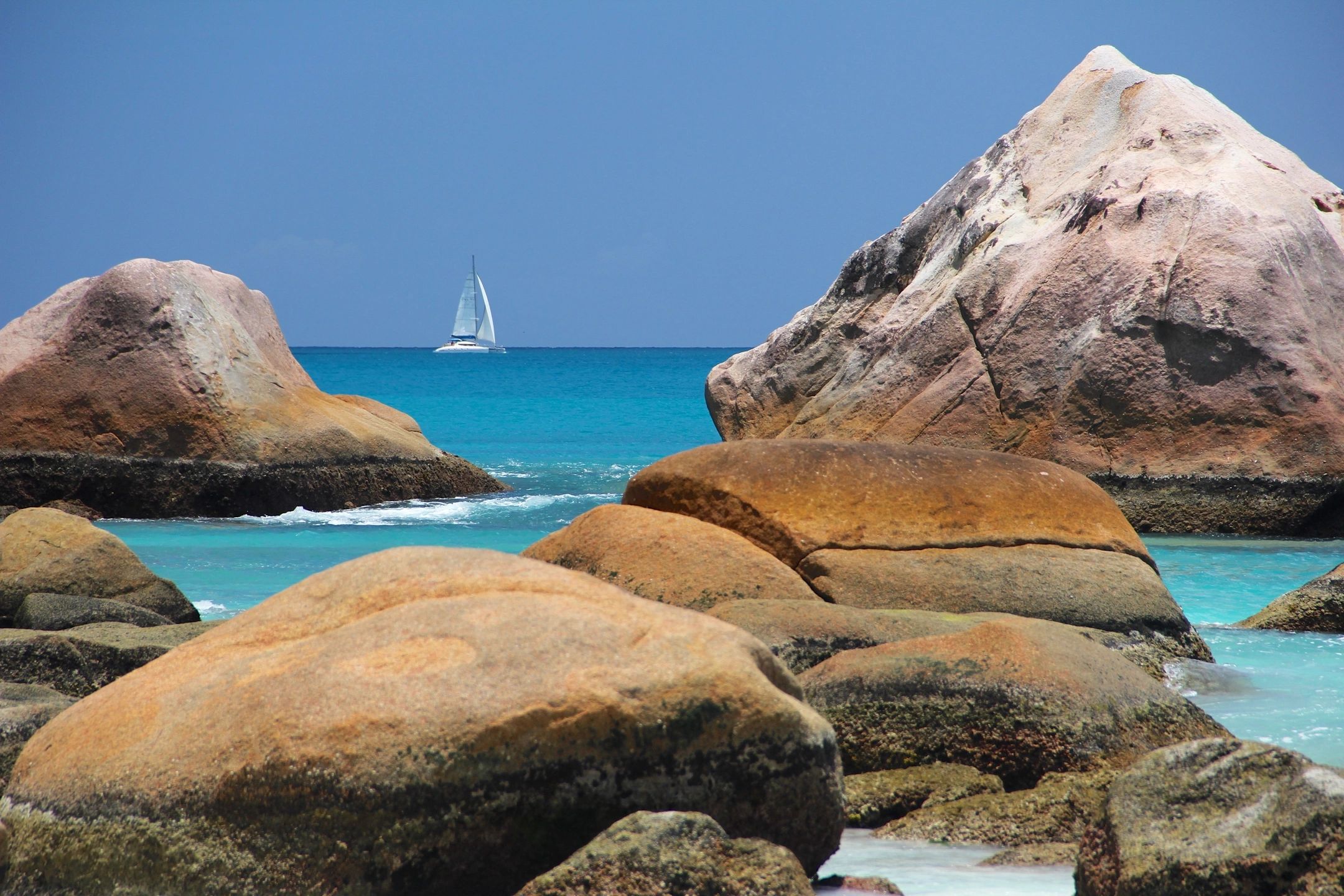 Sailboat sailing on turquoise sea beyond large coastal rocks.