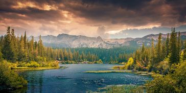 A serene river flows through a lush forest with mountains under a dramatic sunset sky.