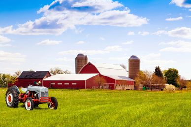 A red tractor on green grass in front of a red barn with silos under a partly cloudy sky.
