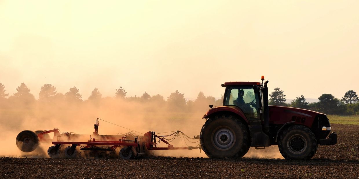 A tractor plowing a dusty field at sunset with trees in the background.