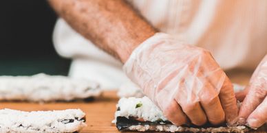 Hands wearing gloves preparing sushi on a wooden board.