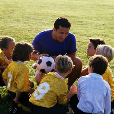 A group of small children are sitting on the grass, listening to a soccer coach who is crouched down