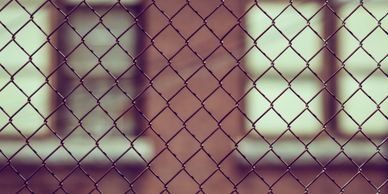 Close-up of a rusty chain-link fence with blurred windows in the background.