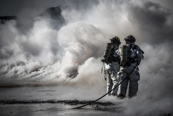 Firefighters in silver suits battle heavy smoke with a hose.