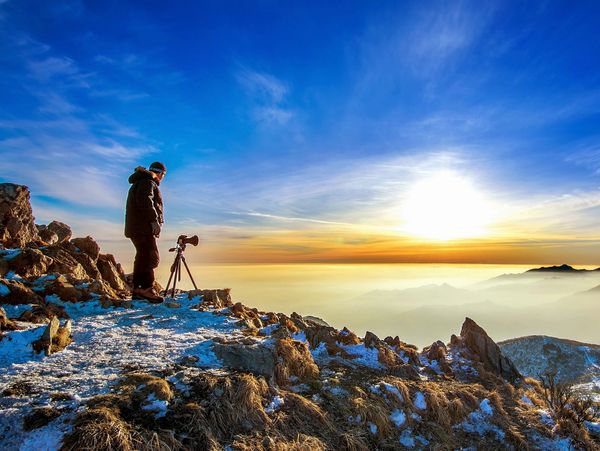 Photographer on rocky mountaintop capturing sunrise over misty mountains.