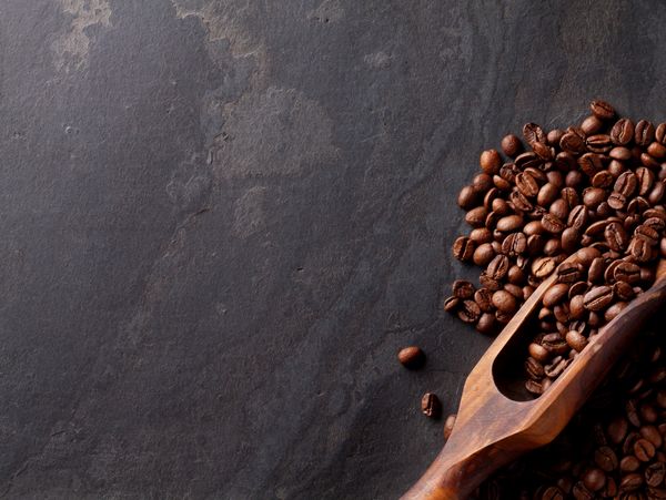 Coffee beans spilling from a wooden scoop on a dark surface.