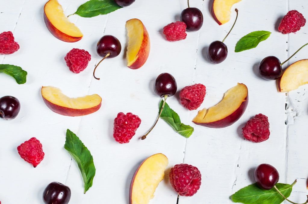 Fresh raspberries, cherries, peach slices, and green leaves on a white wooden surface.
