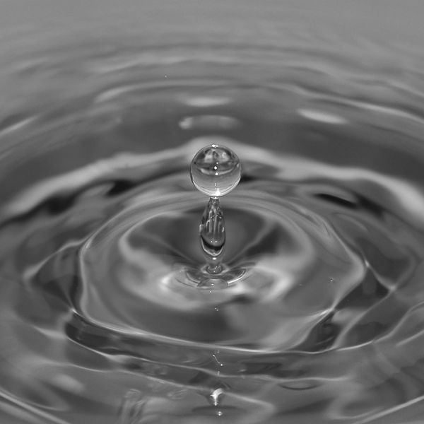 A single water droplet rising from a calm blue surface with ripples.