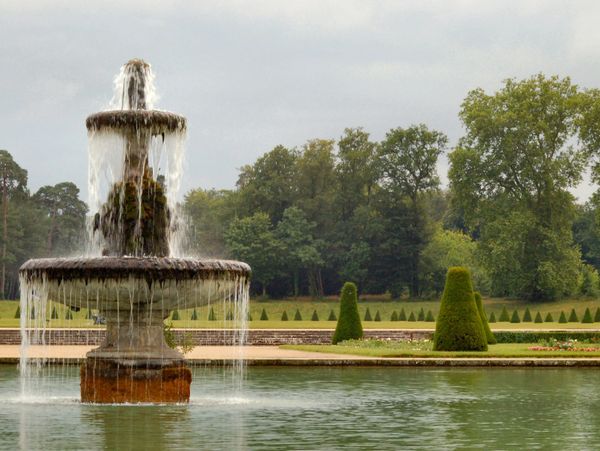 A tiered water fountain in a tranquil garden pond with greenery and trees in the background.