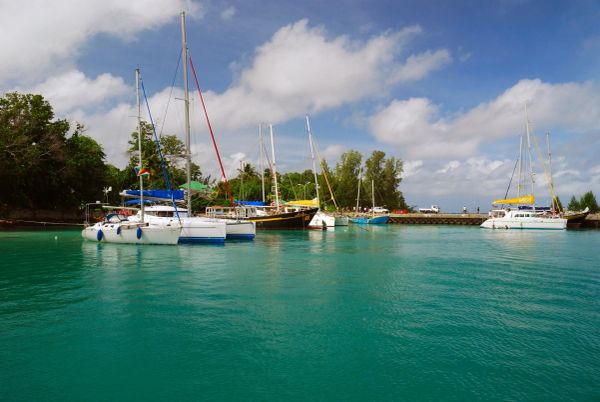 Sailboats docked in calm turquoise waters near a green shoreline under a partly cloudy sky.