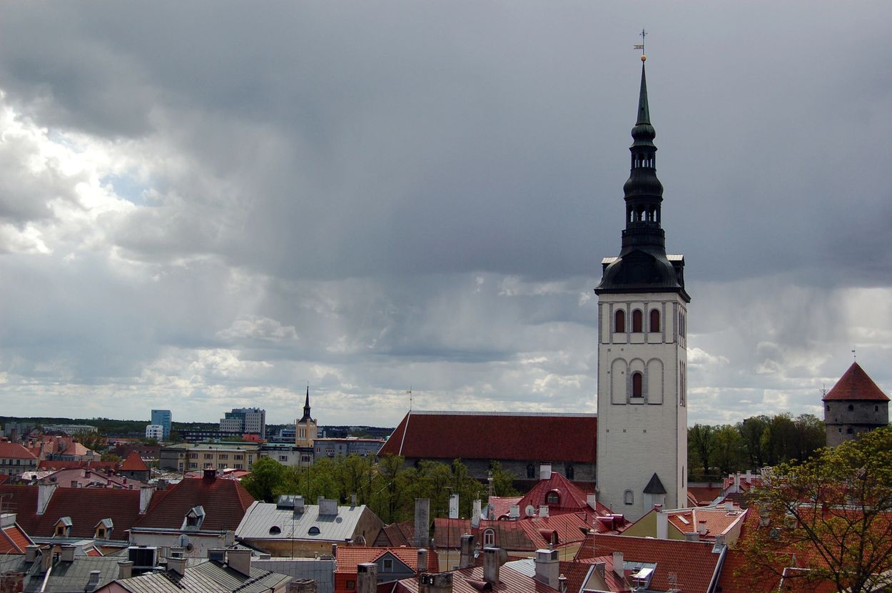 Tall church tower amidst old European rooftops under a cloudy sky.