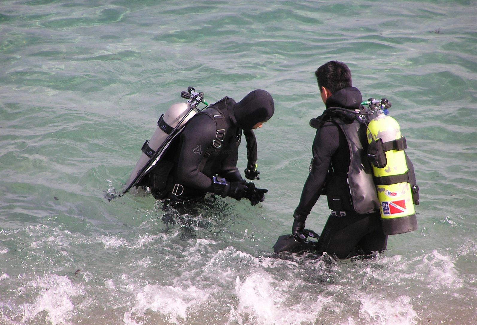 Two scuba divers in wetsuits entering clear ocean water with tanks.