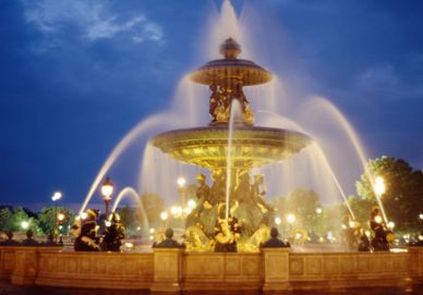 Illuminated ornate fountain with water jets at night under a deep blue sky.