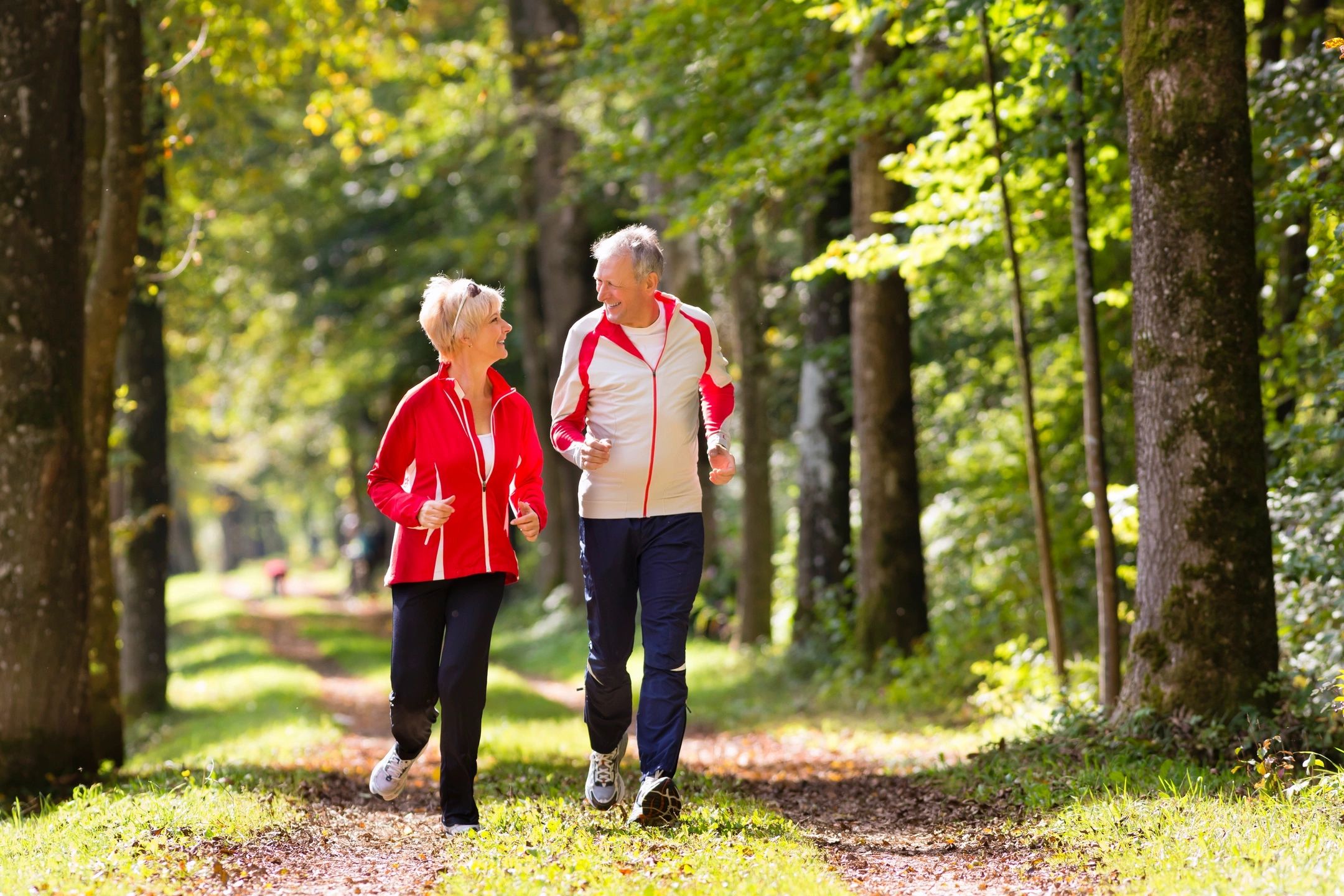 Older couple jogging happily through a sunlit forest path.