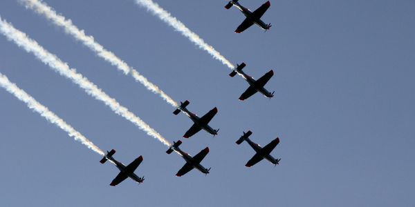 Seven planes flying in formation with smoke trails against a clear blue sky.
