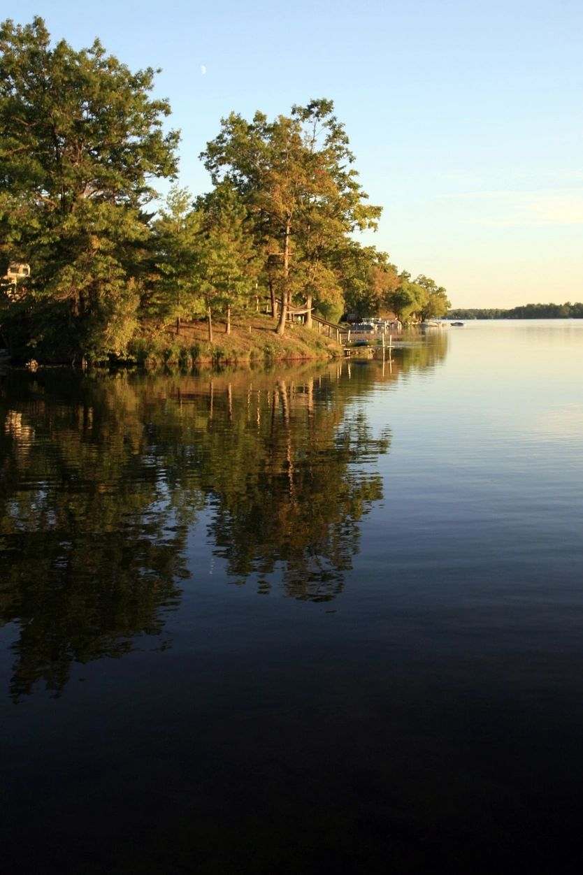 Calm lake with tree-lined shore reflecting in the water at sunset.