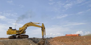 Yellow excavator digging earth with a truck in the background under a blue sky.