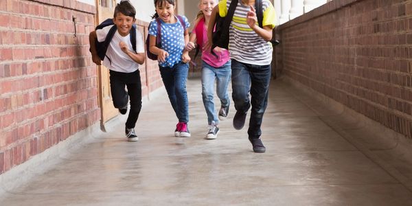 Four children joyfully running down a school hallway with backpacks.