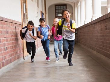 Four children joyfully running down a school corridor with backpacks.