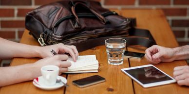 Two people at a table, one taking notes, with a tablet, glass of water, coffee & messenger bag 