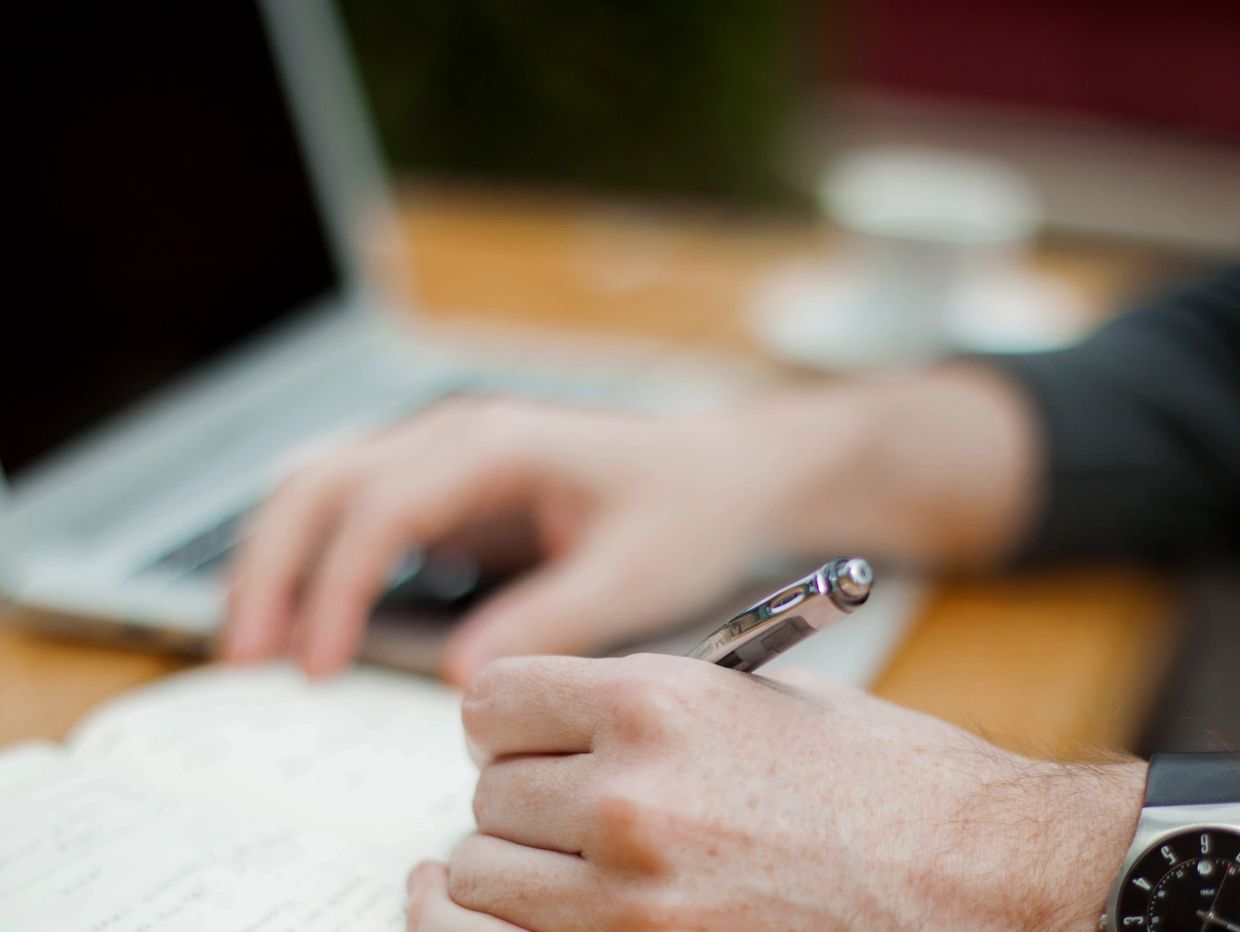 Person writing notes by hand with a laptop in the background.