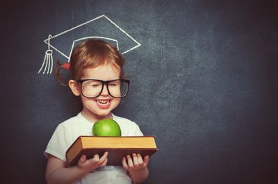 Blackboard with sketch of graduation cap behind the head of smiling child holding  book and apple.