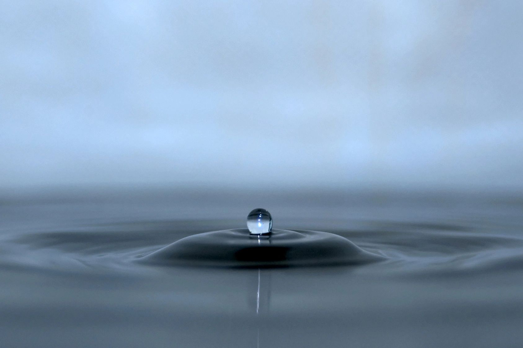 A water droplet captured mid-air above a ripple on a calm surface.