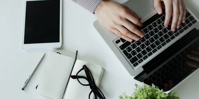 Man typing on a laptop with a tablet, open notebook, glasses, a pen, and a plant on the desk.