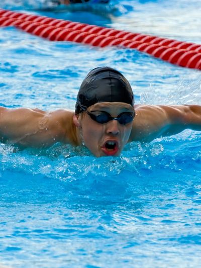 Swimmer in black cap and goggles performing butterfly stroke in pool.