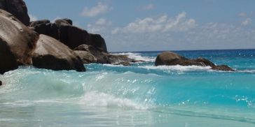 Clear turquoise waves crash gently on rocky beach under blue sky.