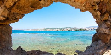 View of clear blue sea and distant town through a rocky cave opening.