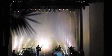 Silhouetted musicians perform on a softly lit stage with a white backdrop and overhead lights.
