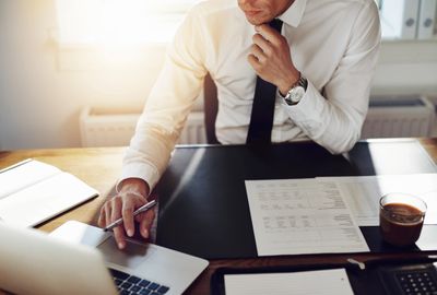 An employee fixing his tie while working on a laptop in his office with coffee on his desk.