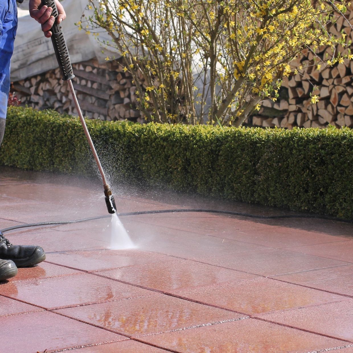 Person pressure washing a brick patio outdoors near shrubbery.