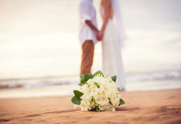 Wedding bouquet on beach with couple holding hands in background.