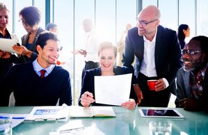 Conference room, people siting at a table and standing, reviewing documents, smiling.