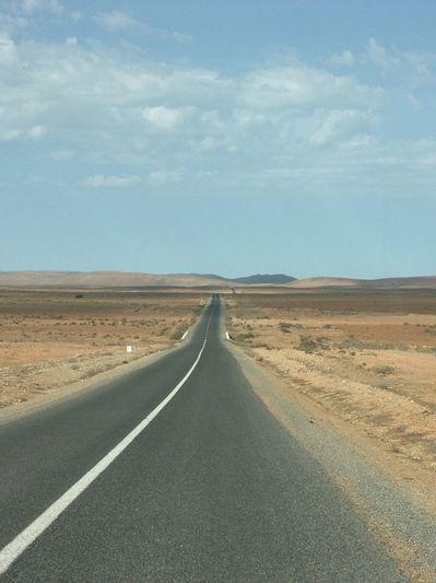 A long straight road stretches through a barren desert landscape under a partly cloudy sky.