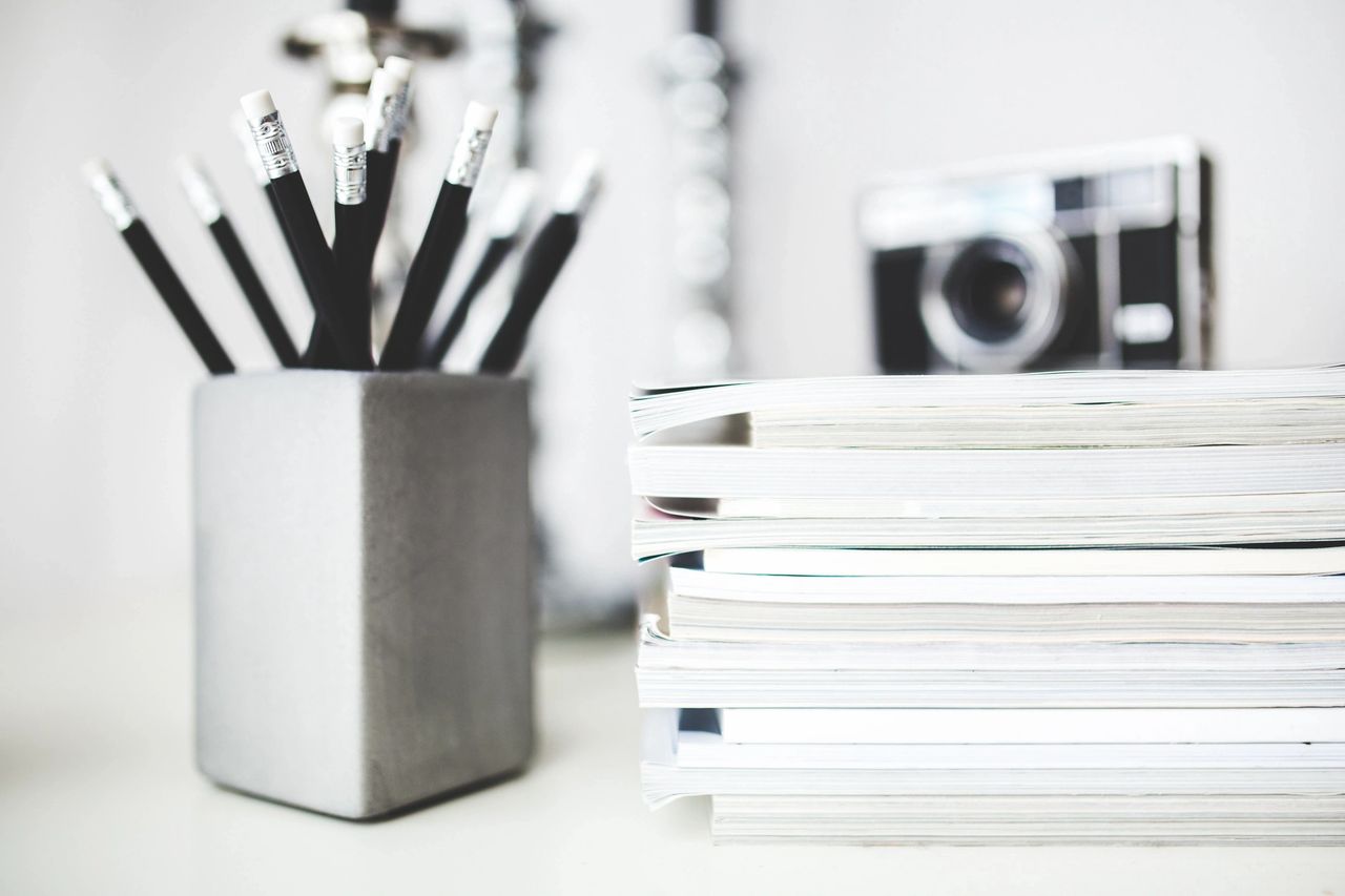 A pencil holder is full of black pencils sat along side a stack of notebooks with a camera on top of them.