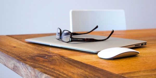 closed laptop with glasses on top and a wireless mouse on brown desk