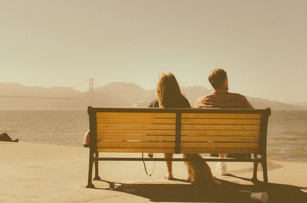 A couple sits on a bench overlooking the water with a distant bridge.