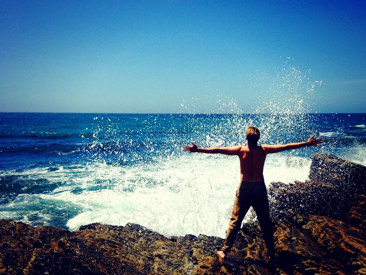 Man standing with open arms on rocks being sprayed by the sea