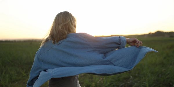 Woman walking away from us in the sunshine up a grassy hill with her right arm outstretched.